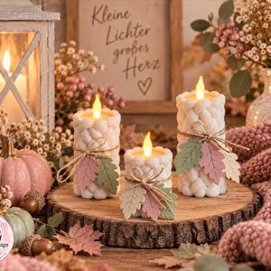 Puede incluir: Tres velas de pilar color crema con un patrón de tejido de punto trenzado texturizado, cada una con una llama LED cálida. Las velas están decoradas con lazos de cuerda y pequeñas hojas de papel multicolor en tonos rosa, verde y beige.