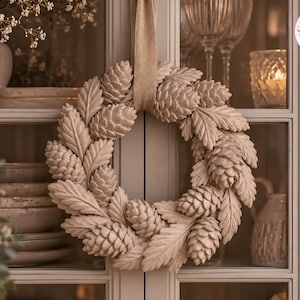 May include: A circular wreath composed of beige, textured pinecones and elongated leaves, suspended by a matching fabric ribbon. The wreath features a monochromatic, matte finish with detailed botanical textures throughout the arrangement.