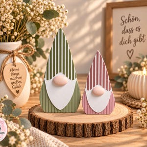Op de afbeelding: Twee houten kabouterfiguren met puntmutsen, één in olijfgroen met witte strepen en één in oudroze met witte strepen. Beide hebben een witte baard en een lichtroze neus. Ze staan op een ronde, natuurlijke houten schijf.