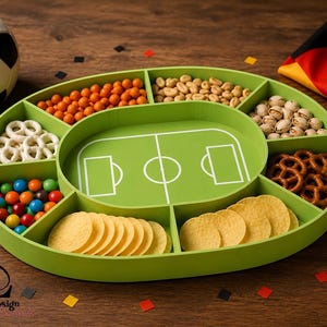 Pode incluir: Uma bandeja de lanches verde oval com compartimentos, com um design de campo de futebol no centro. A bandeja é preenchida com vários lanches, incluindo pretzels, batatas fritas, nozes e doces. Uma bola de futebol e uma bandeira alemã também são visíveis.