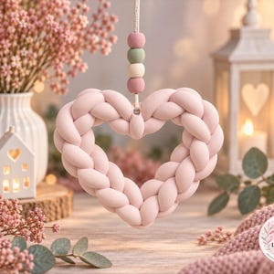 Könnte beinhalten: Ein hängendes herzförmiges Ornament aus geflochtenem hellrosa Stoff. Das Herz hängt an einer Schnur mit vier gestapelten Holzperlen in gedeckten Rosa-, Salbeigrün- und Cremetönen. Das Design ist weich, abgerundet und symmetrisch gestaltet.