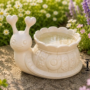 Könnte beinhalten: Ein cremefarbener, schneckenförmiger Keramik-Pflanztopf mit zwei Antennen, die in Herzformen enden, und einem Gehäuse mit gewelltem Rand und geprägten Blumenmustern. Die Schnecke hat eine glatte Oberfläche und einen breiten Boden, mit dem Text Lumito Design.