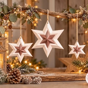 May include: Three hanging star ornaments featuring a white outer frame and a metallic rose gold centre. Each star has a six-pointed geometric design and is suspended by a simple jute twine string from a branch, creating a festive holiday display.