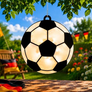 Könnte beinhalten: Eine fußballförmige dekorative Leuchte hängt an einem Ast. Der schwarz-weiße Ball ist beleuchtet, mit deutschen Flaggen und Blumen im Hintergrund. Ein Fußball und eine deutsche Flagge liegen auf einem Holztisch.