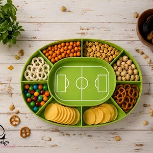 Op de afbeelding: Een ovaal groen serveerblad met een centrale voetbalveld-afbeelding en zes omliggende vakken gevuld met diverse snacks, waaronder krakelingen, noten en kleurrijke snoepjes. De tekst Lunito Design staat linksonder op het dienblad.