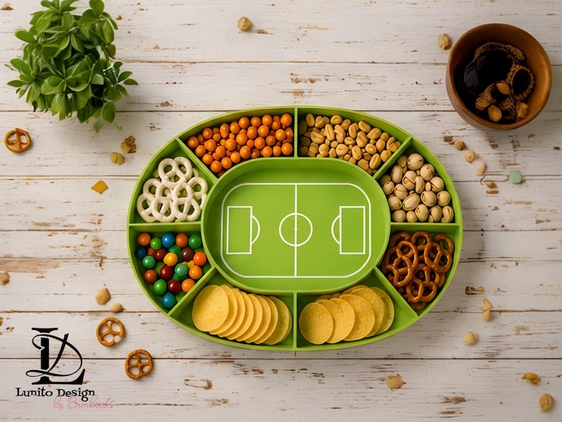 Op de afbeelding: Een ovaal groen serveerblad met een centrale voetbalveld-afbeelding en zes omliggende vakken gevuld met diverse snacks, waaronder krakelingen, noten en kleurrijke snoepjes. De tekst Lunito Design staat linksonder op het dienblad.