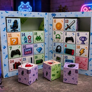 May include: A light blue cardboard advent calendar featuring a pixelated gaming theme with numbered boxes. Each box displays various video game icons including controllers, a sword, a trophy, a headset, and the text GAME OVER and LEVEL.