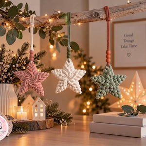 May include: Three macrame star ornaments in dusty pink, cream, and sage green hang from a wooden branch. Each star features a textured, braided cord design, a small wooden bead accent, and a hanging loop. The logo Lunito Design by Benicbooks is visible.