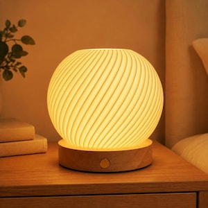 May include: A spherical table lamp featuring a white, ribbed, swirling pattern that emits a warm, soft glow. The lamp is mounted on a circular, light-toned wooden base that includes a small, centered circular power button on the front side.