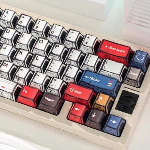 May include: A custom mechanical keyboard with a unique comic book style design. The keycaps are white with black outlines and feature letters, numbers, and symbols. Colored keys include red, blue, and brown. The keyboard has a beige frame.