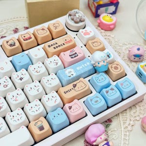May include: A white keyboard with pastel blue, pink, and orange keycaps featuring cute cat illustrations. The keyboard has a 'Backspace' key, an 'Enter' key, and a 'Shift' key.