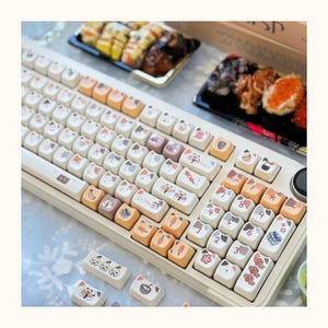 May include: A white mechanical keyboard with custom keycaps featuring cute cartoon animals and food. The keycaps are beige, brown, and pink with black text. The keyboard has a number pad and a function row.