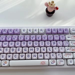 May include: A white keyboard with purple keys and cute cartoon illustrations on the keys. The keyboard has a white space bar with a pink and white illustration. The keyboard is on a white surface.