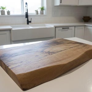May include: A wooden chopping board with a live edge. The board is made of light brown wood with darker brown grain patterns. The board is on a white countertop in a kitchen setting.