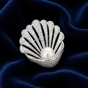 May include: A silver-toned seashell brooch with a central pearl. The brooch is encrusted with small, sparkling stones, and set against a dark blue velvet background. The design is elegant and detailed.