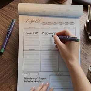 May include: A white weekly planner notepad with black grid lines and text in Turkish, including the word "haftalık" at the top. The planner features sections for days of the week, notes, and next week, with a purple fountain pen resting on the paper.