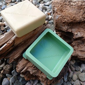 May include: Two square silicone moulds, one beige and one green, sit on wood and pebbles. The beige mould has the text "info@silmoulds.com". The green mould is open, showing its interior. Both moulds have a square shape with a slightly raised edge.