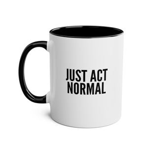 May include: White ceramic coffee mug with a black rim and handle. The mug has the text "JUST ACT NORMAL" printed in black on the front.