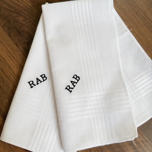 Puede incluir: Tres pañuelos blancos con un sutil patrón de cuadrícula se muestran sobre una superficie de madera. Cada pañuelo está bordado con las letras "RAB" en hilo negro. Un diseño sencillo y elegante.