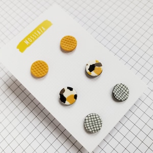 Peut inclure: Ensemble de six petites boucles d'oreilles rondes avec différents motifs. Deux boucles d'oreilles ont un motif gaufré jaune, deux ont un motif quadrillé noir et blanc, et deux ont un motif abstrait noir, blanc et jaune. Les boucles d'oreilles sont présentées sur une carte blanche avec le mot "MOSSTAZA" écrit en jaune.