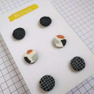 Könnte beinhalten: Fünf runde Keramikohrringe auf einer weißen Karte mit dem in Gelb gedruckten Wort "MOSSAZA". Die Ohrringe sind schwarz, weiß und orange mit geometrischen Mustern.