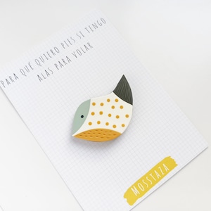 Puede incluir: Un pin de pescado blanco, amarillo y gris con lunares negros y blancos. El pin dice "PARA QUE QUIERO PIES SI TENGO ALAS PARA VOLAR" y "MOSSTAZA" en letras amarillas.