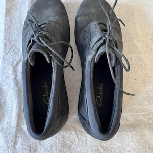 Op de afbeelding: Een paar donkergrijze Clarks veterschoenen. De schoenen hebben een textuur en zichtbare veters. De binnenkant van elke schoen heeft de merknaam "Clarks" in gouden letters gedrukt. De schoenen worden gepresenteerd op een lichtgekleurde stof.