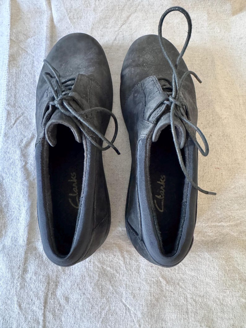 Op de afbeelding: Een paar donkergrijze Clarks veterschoenen. De schoenen hebben een textuur en zichtbare veters. De binnenkant van elke schoen heeft de merknaam "Clarks" in gouden letters gedrukt. De schoenen worden gepresenteerd op een lichtgekleurde stof.