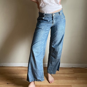 Puede incluir: Pantalones vaqueros de pierna ancha en azul claro con cierre de botón. Los pantalones se combinan con una camiseta blanca. La modelo está de pie con las manos en las caderas. Los pantalones tienen un estilo clásico y casual.