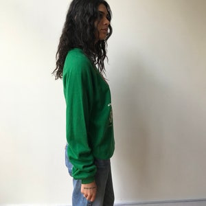 Puede incluir: Una sudadera verde de manga larga con un estampado gr&aacute;fico en el pecho que incluye texto en blanco y dorado. La sudadera tiene cuello redondo y pu&ntilde;os acanalados, combinada con unos vaqueros azules de corte recto y dise&ntilde;o cl&aacute;sico.