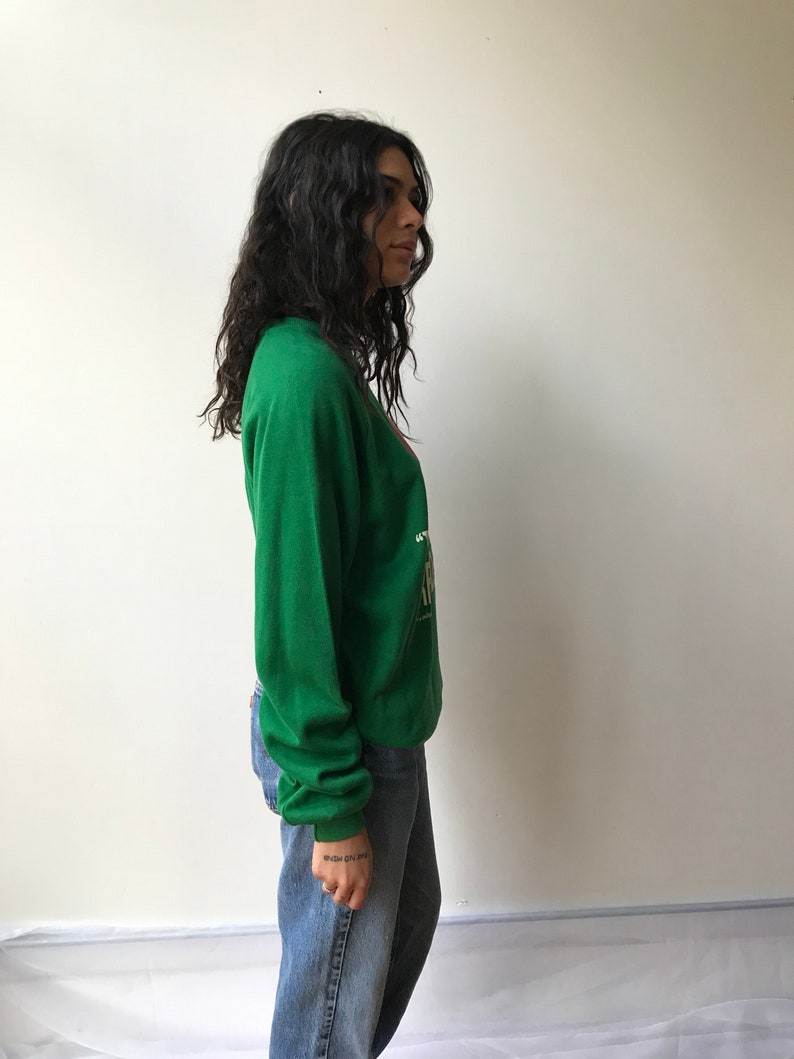 Puede incluir: Una sudadera verde de manga larga con un estampado gr&aacute;fico en el pecho que incluye texto en blanco y dorado. La sudadera tiene cuello redondo y pu&ntilde;os acanalados, combinada con unos vaqueros azules de corte recto y dise&ntilde;o cl&aacute;sico.