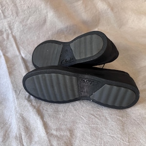Op de afbeelding: Een paar zwarte Clarks-schoenen, gezien vanaf de zolen. De zolen zijn grijs met een textuurpatroon en de merknaam "Clarks" is zichtbaar. De schoenen zijn gemaakt van een donker materiaal en staan tegen een neutrale achtergrond.