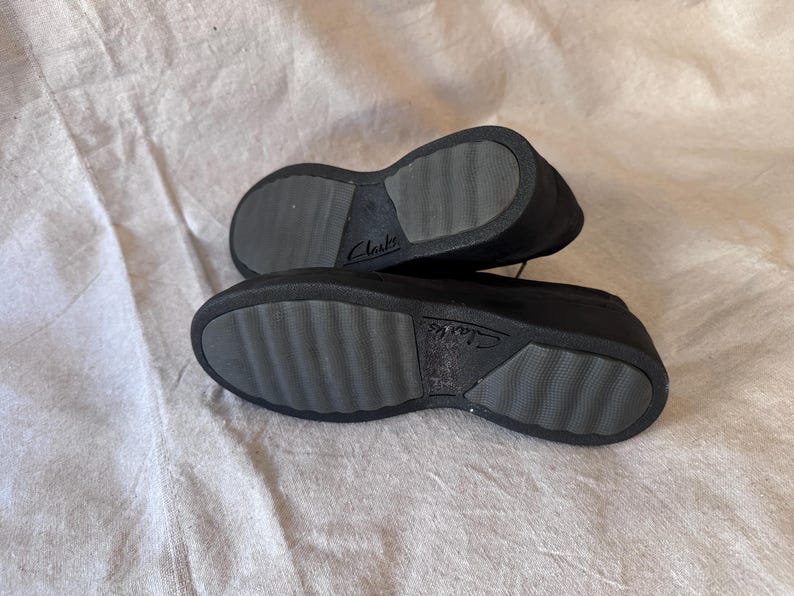Op de afbeelding: Een paar zwarte Clarks-schoenen, gezien vanaf de zolen. De zolen zijn grijs met een textuurpatroon en de merknaam "Clarks" is zichtbaar. De schoenen zijn gemaakt van een donker materiaal en staan tegen een neutrale achtergrond.