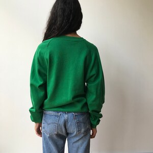 Puede incluir: Una sudadera de color verde s&oacute;lido con cuello redondo y mangas largas combinada con pantalones vaqueros de mezclilla azul claro. La sudadera presenta un dise&ntilde;o sencillo con pu&ntilde;os y cuello acanalados, mientras que los vaqueros tienen un corte cl&aacute;sico.
