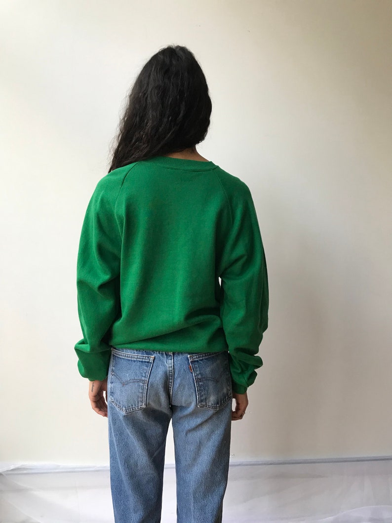 Puede incluir: Una sudadera de color verde s&oacute;lido con cuello redondo y mangas largas combinada con pantalones vaqueros de mezclilla azul claro. La sudadera presenta un dise&ntilde;o sencillo con pu&ntilde;os y cuello acanalados, mientras que los vaqueros tienen un corte cl&aacute;sico.