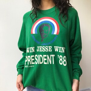Puede incluir: Una sudadera verde de manga larga con un gr&aacute;fico de Jesse Jackson bajo un arco&iacute;ris rojo, blanco y azul. El texto dice WIN JESSE WIN, PRESIDENT '88 y W.C. enterprises. El dise&ntilde;o est&aacute; impreso en tinta blanca, azul y roja en el frente.