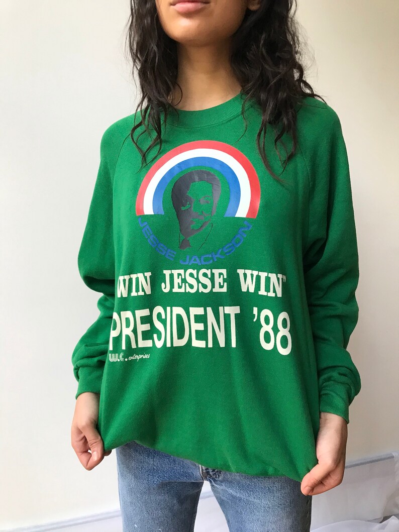 Puede incluir: Una sudadera verde de manga larga con un gr&aacute;fico de Jesse Jackson bajo un arco&iacute;ris rojo, blanco y azul. El texto dice WIN JESSE WIN, PRESIDENT '88 y W.C. enterprises. El dise&ntilde;o est&aacute; impreso en tinta blanca, azul y roja en el frente.