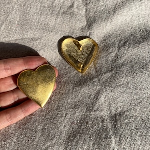 May include: Two gold-colored heart-shaped brooches. One brooch is held in a hand, while the other lies on a textured, light-colored surface. The brooches have a smooth, reflective surface, and the lighting creates shadows.