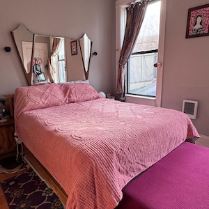 May include: A bedroom featuring a bed with a pink textured quilt. The bed has a wooden frame and a magenta bench at the foot. A decorative mirror headboard and a patterned rug are also visible.