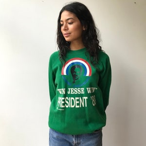 Puede incluir: Una sudadera verde de cuello redondo con un gr&aacute;fico de Jesse Jackson bajo un arco&iacute;ris rojo, blanco y azul. El texto dice "WIN JESSE WIN" y "PRESIDENT '88" en letras blancas. La sudadera se combina con unos pantalones vaqueros azules.