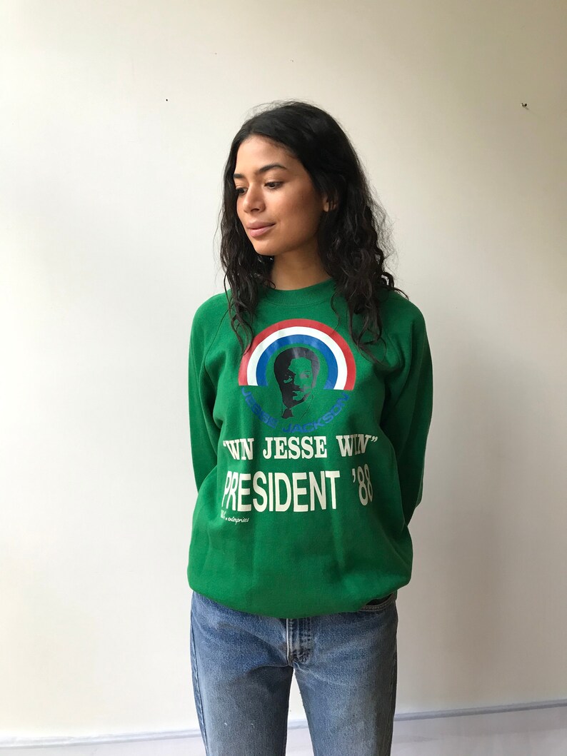 Puede incluir: Una sudadera verde de cuello redondo con un gr&aacute;fico de Jesse Jackson bajo un arco&iacute;ris rojo, blanco y azul. El texto dice "WIN JESSE WIN" y "PRESIDENT '88" en letras blancas. La sudadera se combina con unos pantalones vaqueros azules.