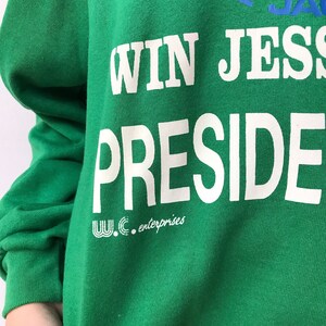 Puede incluir: Una sudadera verde brillante de manga larga con texto impreso en blanco que dice WIN JESS PRESIDENT. Debajo del texto hay un peque&ntilde;o logotipo blanco y las palabras W.C. enterprises. La parte superior incluye texto azul parcialmente visible.