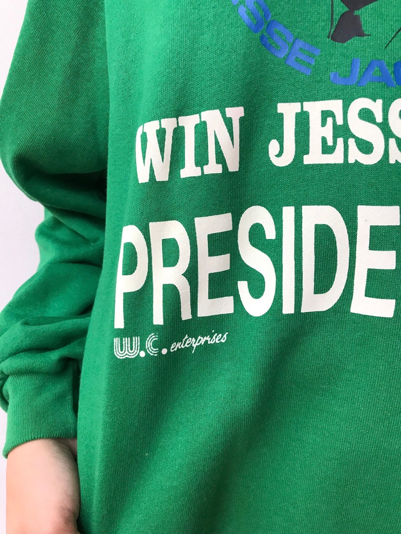Puede incluir: Una sudadera verde brillante de manga larga con texto impreso en blanco que dice WIN JESS PRESIDENT. Debajo del texto hay un peque&ntilde;o logotipo blanco y las palabras W.C. enterprises. La parte superior incluye texto azul parcialmente visible.