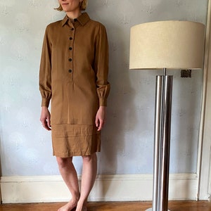 May include: A long-sleeved, knee-length brown dress with a collared neckline and button-down front. The dress features a pleated detail at the hem. A chrome floor lamp with a beige lampshade stands nearby.