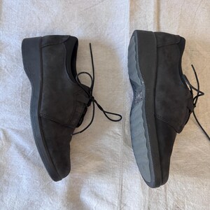 Op de afbeelding: Twee zwarte leren schoenen met veters. De schoenen hebben een lage hak en een bovenwerk met textuur. De veters zijn gestrikt. De schoenen liggen op een witte ondergrond.