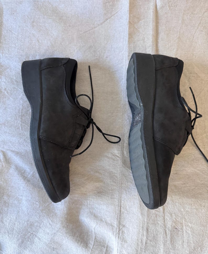 Op de afbeelding: Twee zwarte leren schoenen met veters. De schoenen hebben een lage hak en een bovenwerk met textuur. De veters zijn gestrikt. De schoenen liggen op een witte ondergrond.