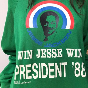 Puede incluir: Una sudadera verde de manga larga con un gr&aacute;fico de Jesse Jackson bajo un arco&iacute;ris rojo, blanco y azul. El texto dice "JESSE JACKSON", "WIN JESSE WIN", "PRESIDENT '88" y "W.C. enterprises" en letras de color blanco y azul.