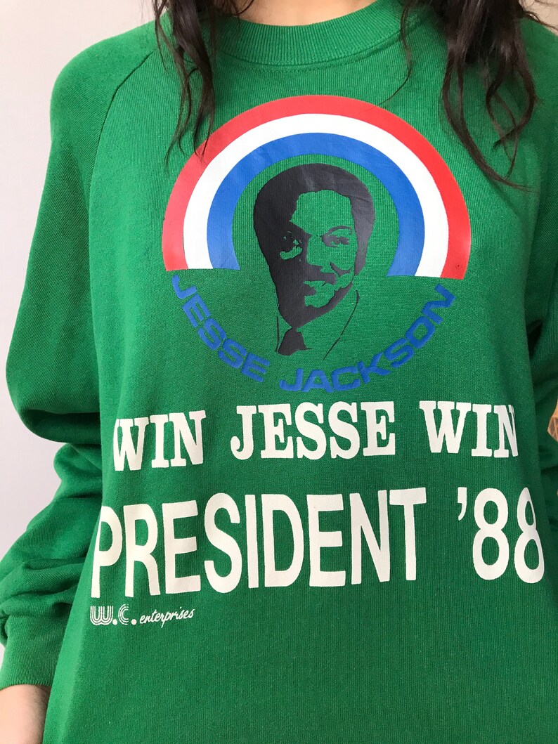 Puede incluir: Una sudadera verde de manga larga con un gr&aacute;fico de Jesse Jackson bajo un arco&iacute;ris rojo, blanco y azul. El texto dice "JESSE JACKSON", "WIN JESSE WIN", "PRESIDENT '88" y "W.C. enterprises" en letras de color blanco y azul.