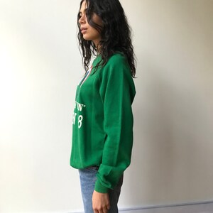 Puede incluir: Una sudadera verde de manga larga con cuello redondo que presenta texto blanco con las palabras "IN" y "8" junto a un elemento gr&aacute;fico parcial en rojo y azul. La sudadera se combina con vaqueros azules, mostrando un ajuste casual y tela verde.