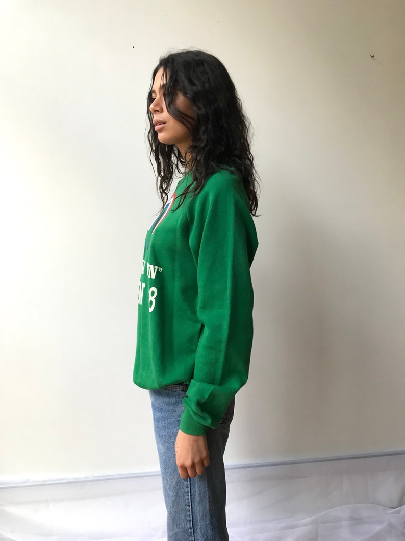 Puede incluir: Una sudadera verde de manga larga con cuello redondo que presenta texto blanco con las palabras "IN" y "8" junto a un elemento gr&aacute;fico parcial en rojo y azul. La sudadera se combina con vaqueros azules, mostrando un ajuste casual y tela verde.